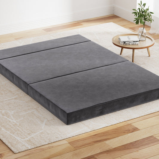 Foldable grey foam mattress laid out on the floor in a room with a small round table and plant to the side.