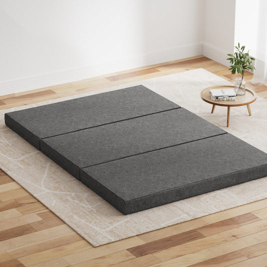 Foldable grey foam mattress laid out on the floor in a room with a small round table and a plant.