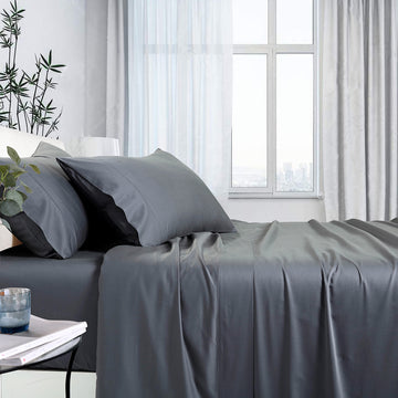 A double-sized sheet set in charcoal color, displayed on a bed with pillows and a flat sheet, in a room with a window and a side table with a plant.