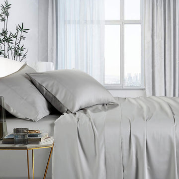 A king-sized silver sheet set on a bed, with a fitted sheet, flat sheet, and pillowcases displayed in a room with a window and a side table with a plant.