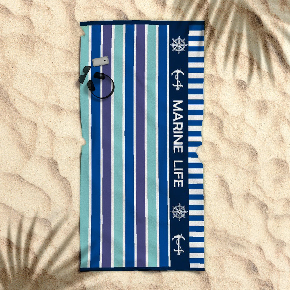 A striped beach towel with marine life graphics, laid out on a sandy surface with a palm leaf shadow over it.