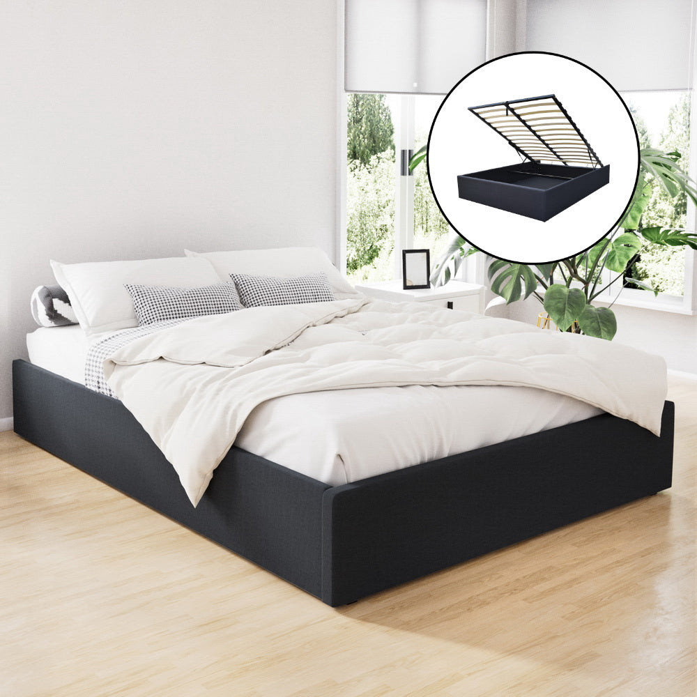 A queen-sized, charcoal-colored gas lift bed frame with a storage compartment underneath, depicted in a room setting with a white bedsheet and pillows on top.