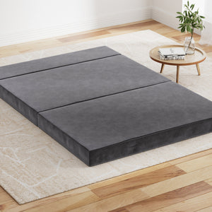 Foldable grey foam mattress laid out on the floor in a room with a small round table and plant to the side.