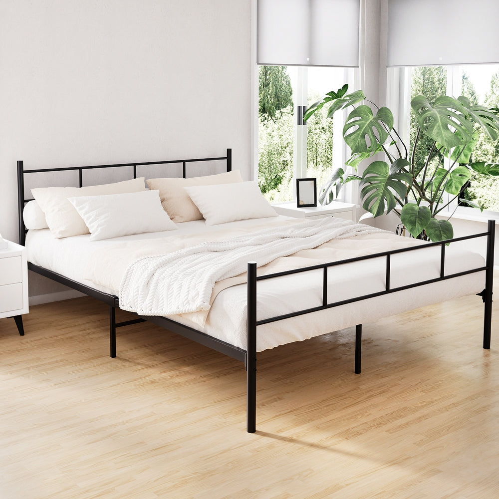 Black metal queen bed frame with a solid headboard and footboard, placed in a room with a white mattress, pillows, and a plant in the background.