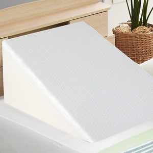 A white, angled wedge pillow made of memory foam, placed on a bed next to a basket with a plant.