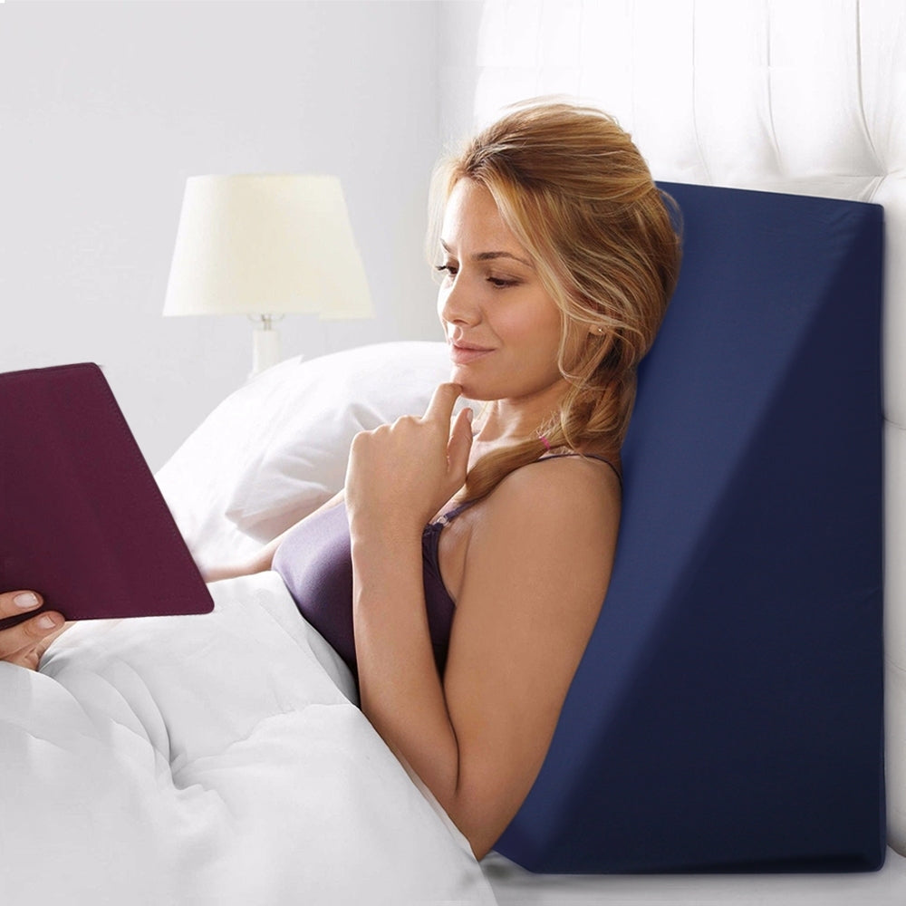 A woman lying on a blue bed wedge pillow while reading a book in bed.