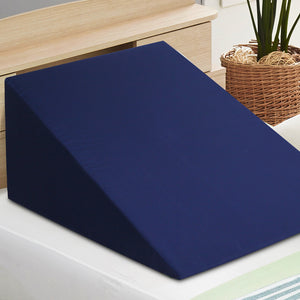 A blue wedge pillow made of memory foam, placed on a bed against a white pillow, with a wooden headboard and a plant in the background.