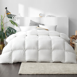 A super king-sized white duck down feather quilt on a bed, with a white cotton cover and a 233 thread count, displayed in a bright bedroom setting with decorative pillows, a bedside lamp, and houseplants.
