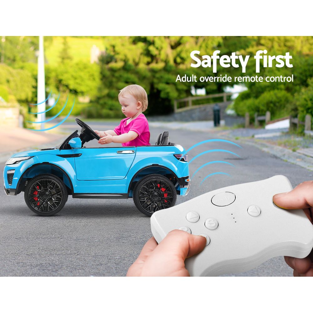Child in a blue toy car with an adult override remote control, outdoors.