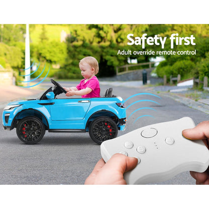 Child in a blue toy car with an adult override remote control, outdoors.