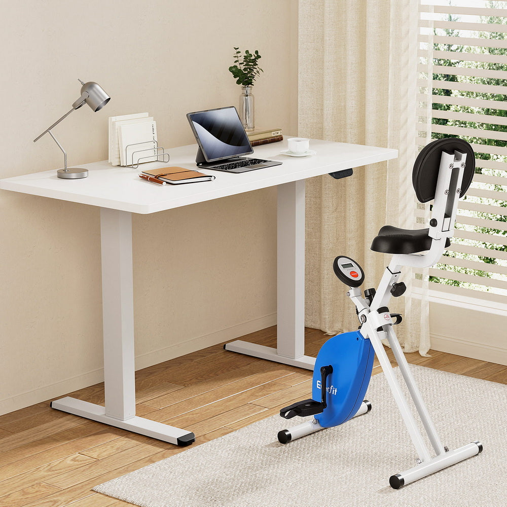 White desk with laptop and stationery items next to a small exercise bike in a room with a window and rug.