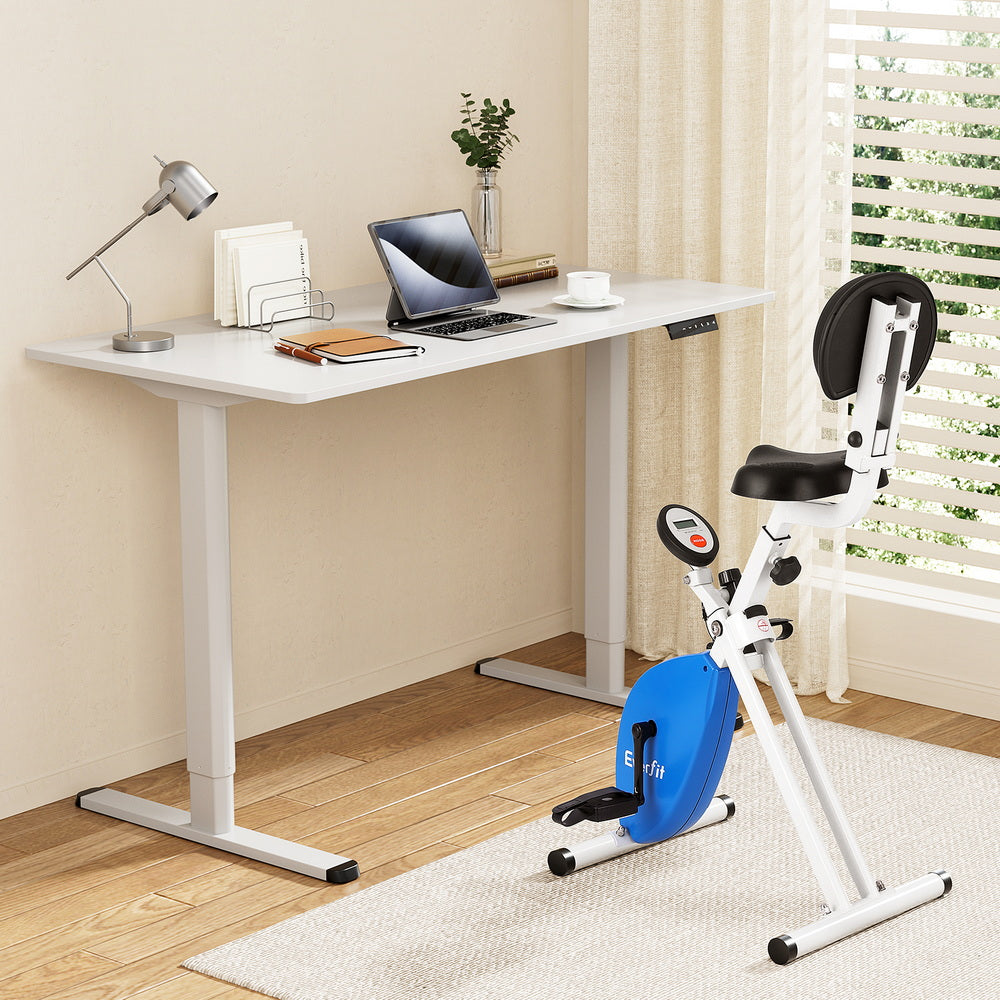 White desk with laptop and books, blue exercise bike in a room with a window.