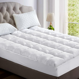 A white mattress topper on a queen-sized bed, with a pillow on top of the topper. The bed is set in a room with a window in the background.