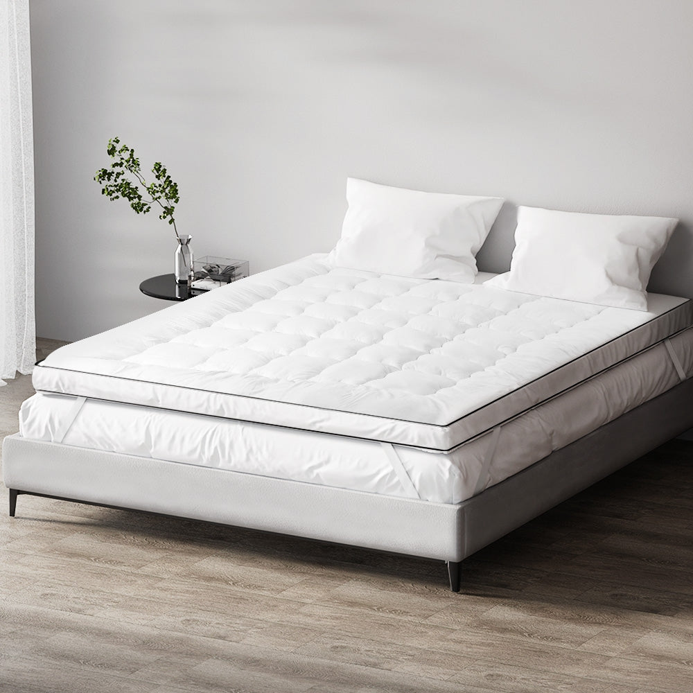 A queen-sized white mattress topper on a bed with a white bedsheet, pillows, and a plant beside the bed.