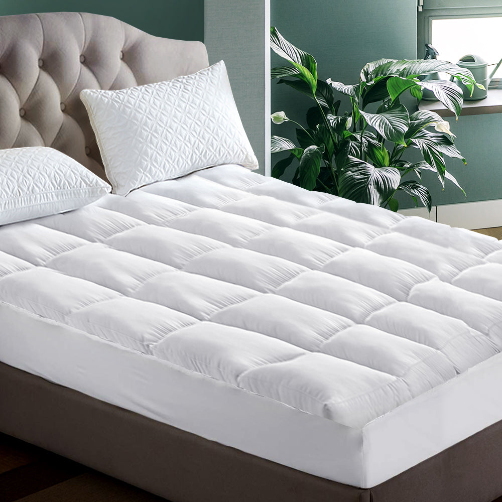 A single-sized mattress topped with a white pillowtop mattress topper, placed on a bed with a beige headboard, in a room with a window and a plant.