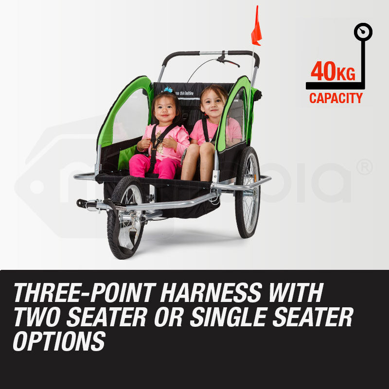 Child bike trailer with two children in pink outfits on a white background, featuring text about capacity and harness options.