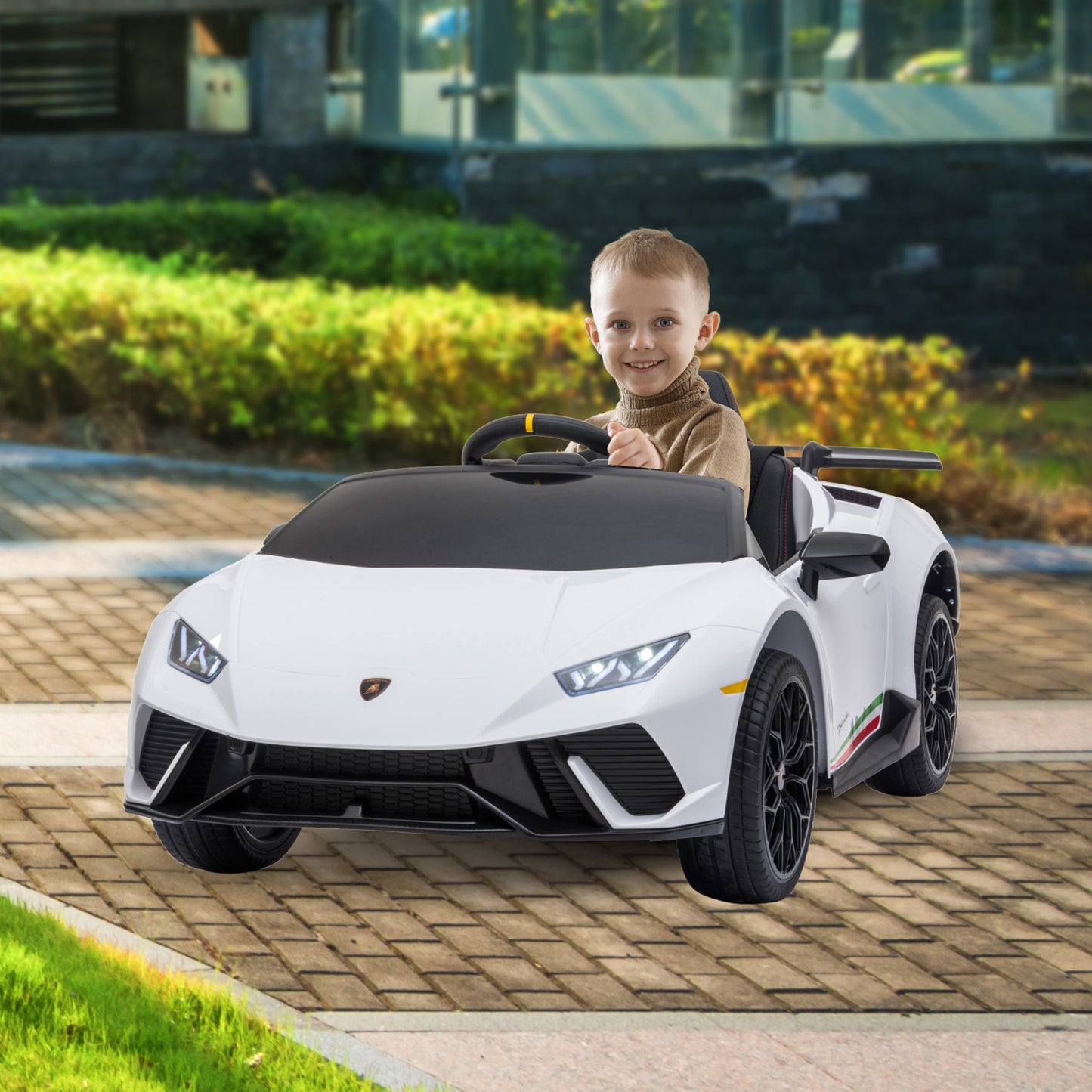 Child sitting in a toy sports car on a paved area with greenery in the background