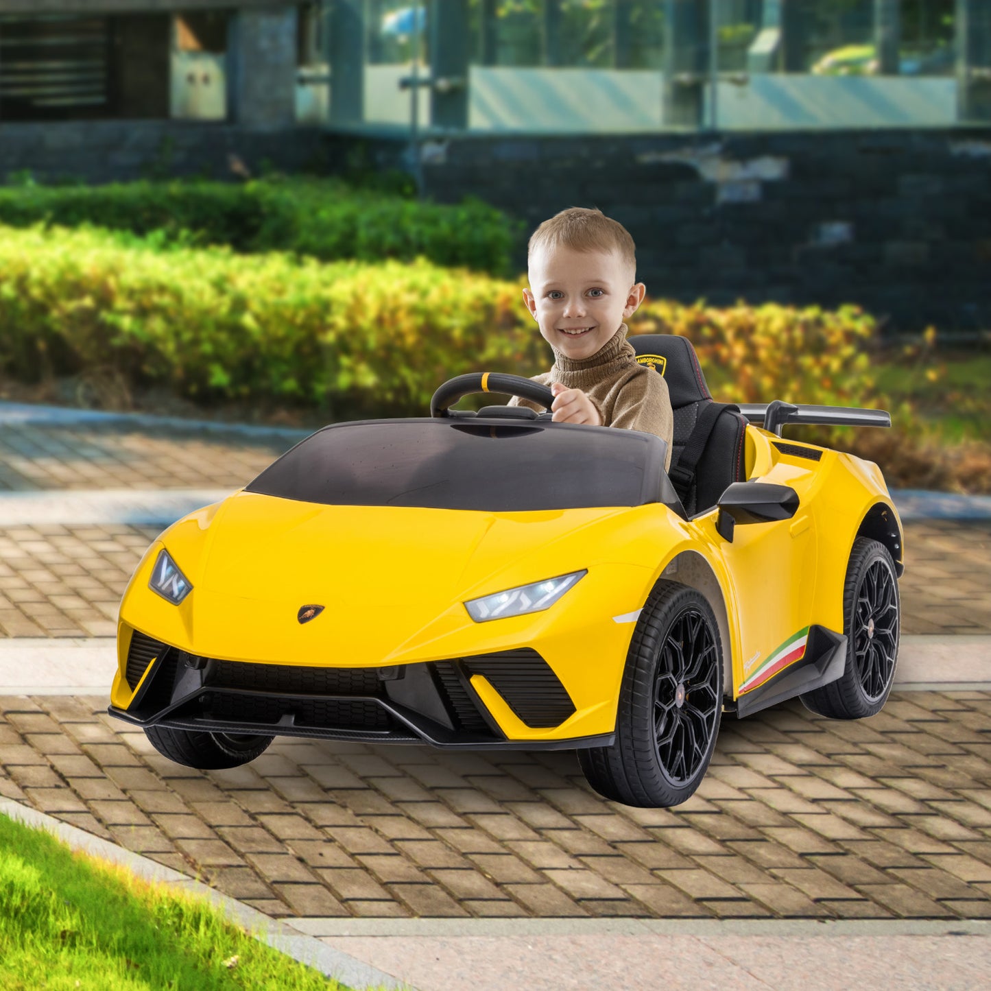 Child sitting in a yellow toy car on a paved area with greenery in the background