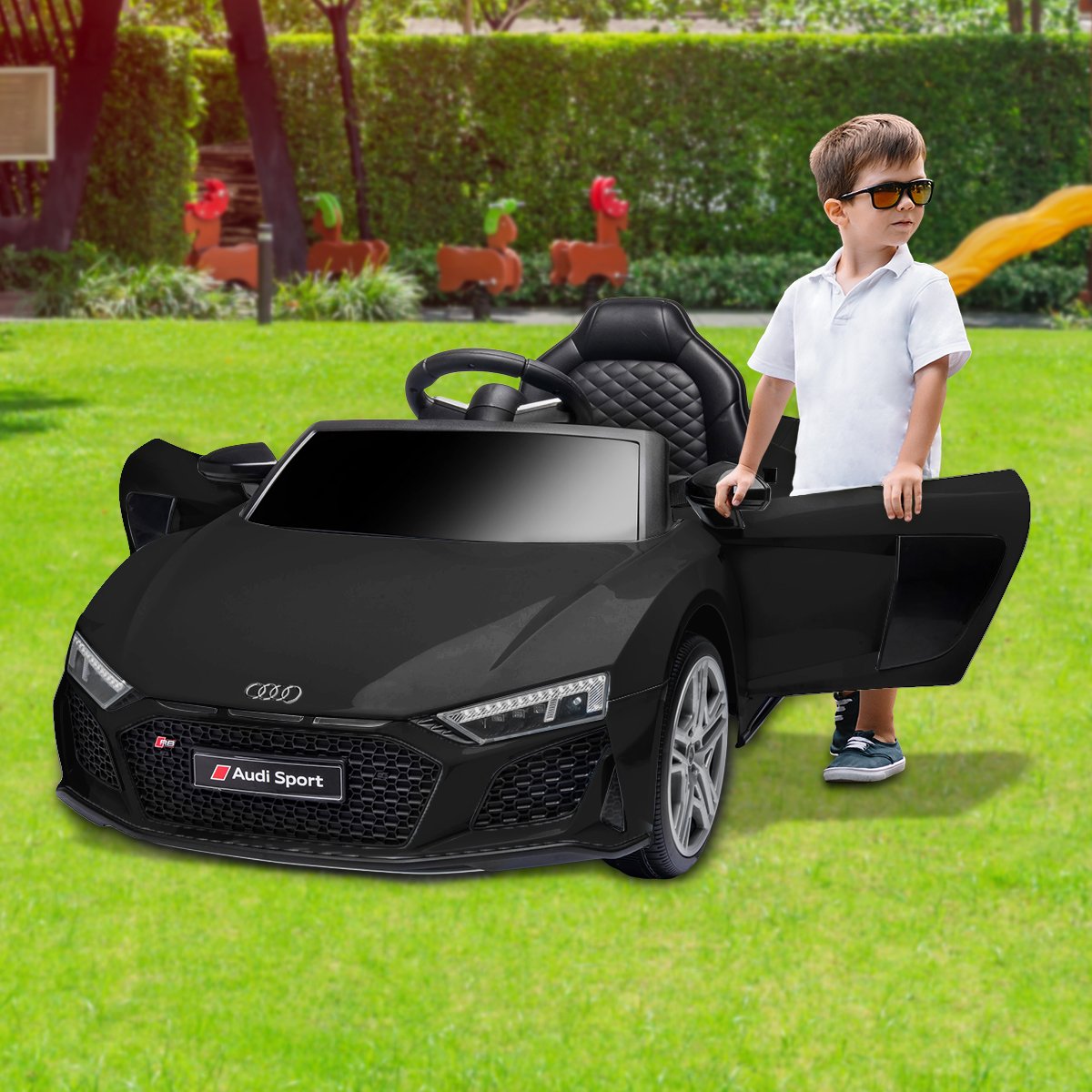 Child standing next to a black Audi toy car on a grassy area with playground equipment in the background.