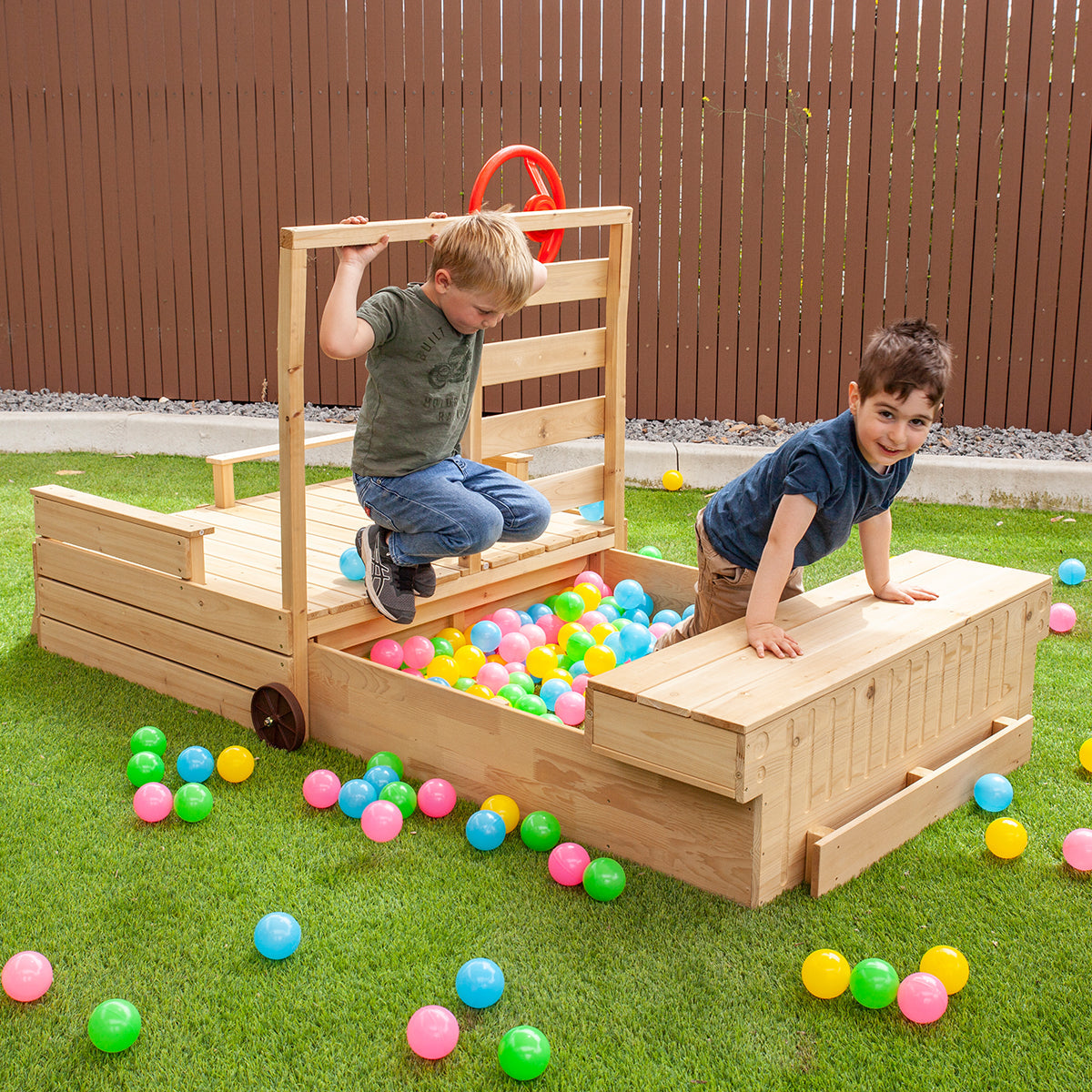 Two children playing with a wooden toy truck filled with colorful balls on a grassy area.