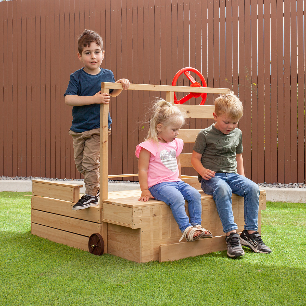 Three children playing on a wooden playground structure with a brown fence and grass background.
