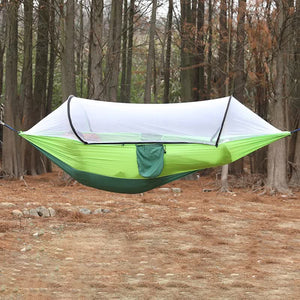 A green outdoor camping hammock with a built-in mosquito net is displayed in a forest setting. The hammock appears to be in use, showing a sense of comfort and protection from insects.