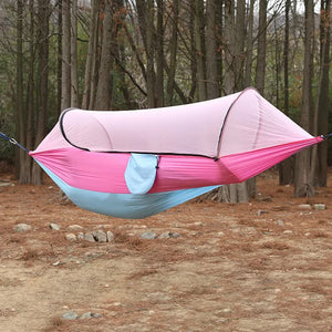 A pink and blue outdoor camping hammock with a built-in mosquito net is set up in a forested area, providing a comfortable-looking resting spot.
