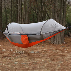 A red and gray outdoor camping hammock with a built-in mosquito net is displayed in a forest setting.