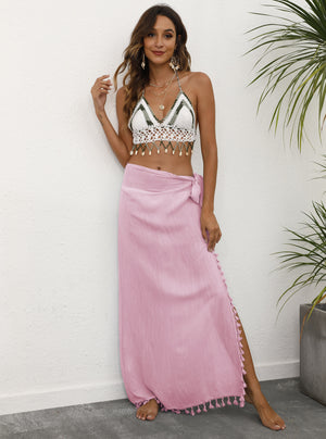 A woman wearing a pink lightweight tassel sarong with tie details at the waist, modeled as a skirt. She is standing against a backdrop with greenery.