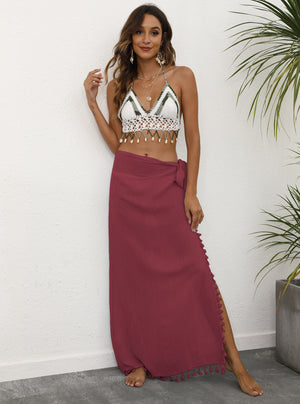 A woman wearing a red lightweight tassel sarong with tie details at the waist and a side slit, paired with a white and gold bikini top.