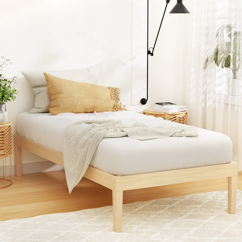 A single-sized wooden bed frame made of pine with a white mattress on it, positioned in a minimally decorated room.