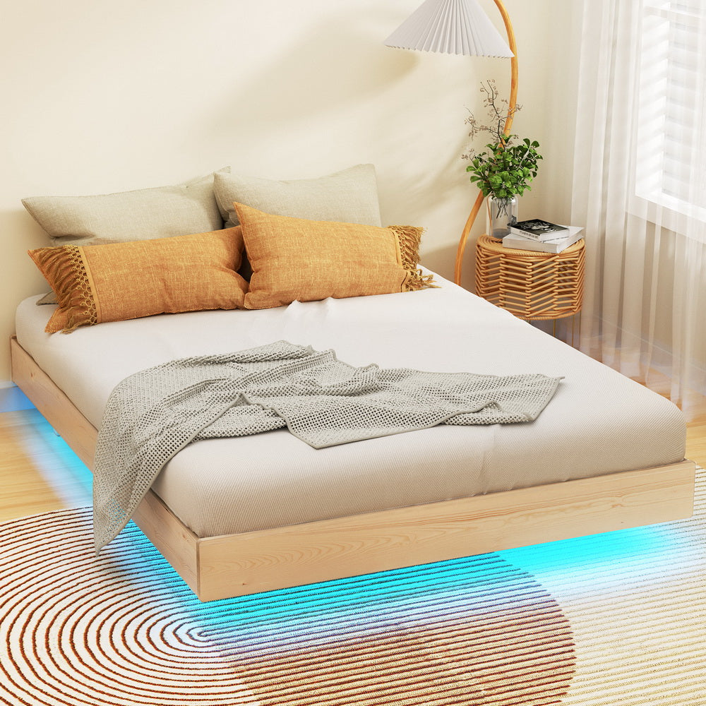 Double size wooden bed frame with a floating design, featuring LED lights under the frame, placed in a room with a white mattress, orange pillows, and a lamp in the background.