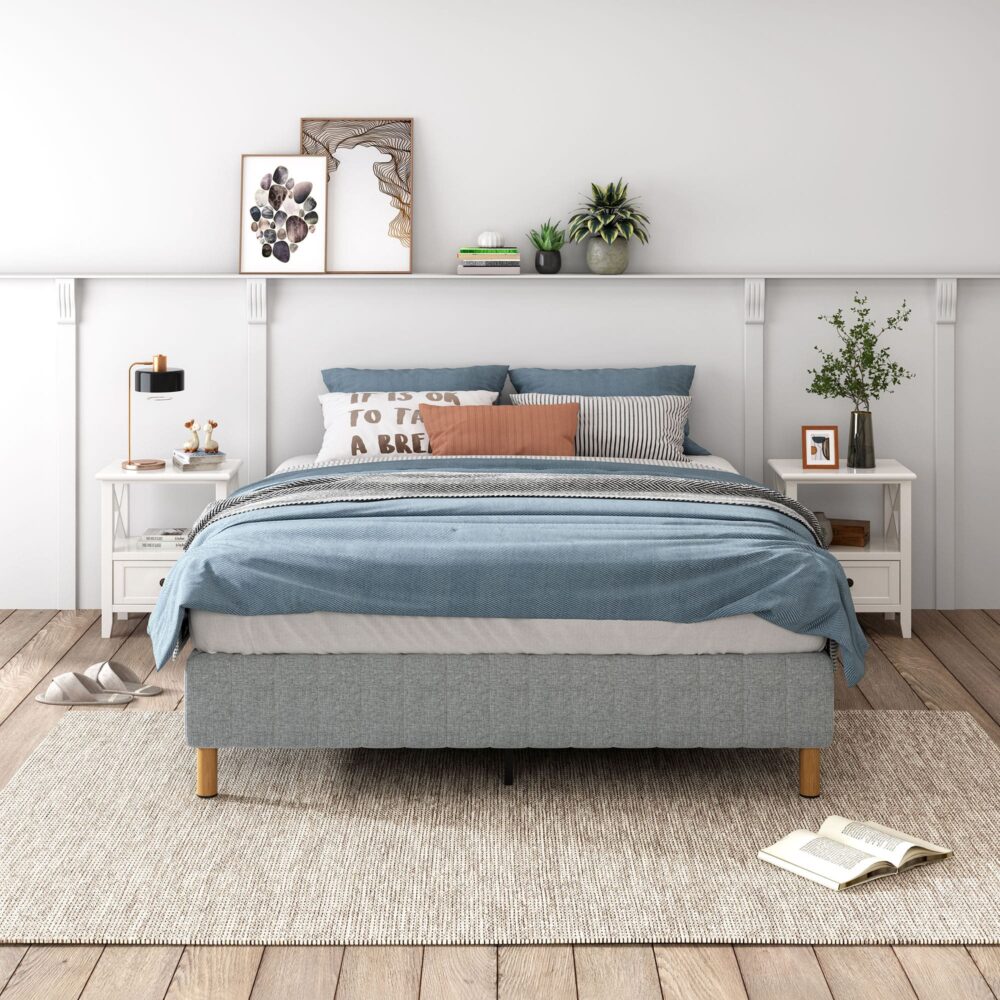 A double-sized metal bed frame with a light grey mattress foundation, covered with a linen fabric. The bed is set in a room with wooden flooring, with a nightstand and a plant to the side, and a framed picture on the wall above.