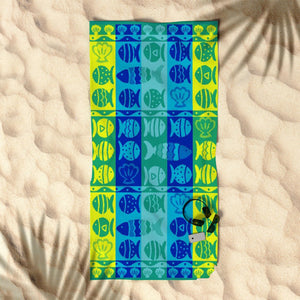 A beach towel with a jacquard nautical design featuring blue, yellow, green, and white colors, laid out on a sandy surface with palm trees in the background.