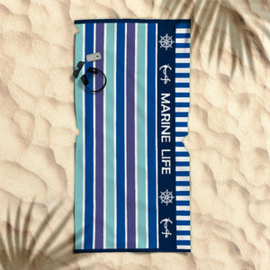 A striped beach towel with marine life graphics, laid out on a sandy surface with a palm leaf shadow over it.