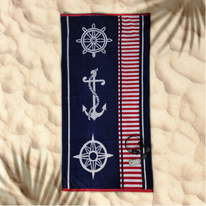 A navy blue beach towel with a nautical jacquard design featuring a anchor, compass, and steering wheel, laid out on a sandy surface.